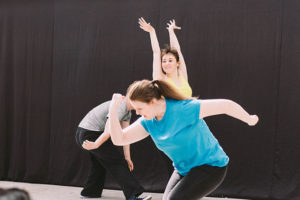 Photo: Three young white people are dancing. The one in front is making a strong-arm fist while slightly bent forward, wearing a blue t shirt. The one in the back has both arms up and fingers spread out. One at the side can only partially be seen.