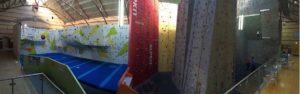 Photo: a panoramic view inside Nottingham Climbing Centre. In the middle part of the photo are various different climbing walls, with handholds dotted over them. On the right is a low balcony area with tables and chairs. On the left in the far distance, a door can be seen. Although the sign on it is too far away to be seen, it's a door to some toilets.