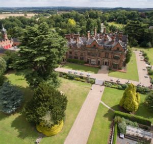 Photo: a large red-brick hall with pointy roofs and tall chimneys is viewed from above, as if from an aeroplane. It stands within formal lawns and is surrounded by woodland. A further courtyard can be glimpsed to the left of the picture. The horizon can be seen in the distance.