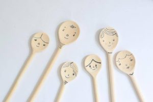 Six wooden spoons each have a smiley face drawn on their round end, and are arranged on a white background like a family of smiling spoons.