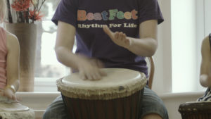 A child's hands can be seen playing an African-style drum. Part of the child's t shirt can be seen. The t shirt has in colourful letters "BeatFeet". Below the "BeatFeet" logo is the phrase "Rhythm For Life".
